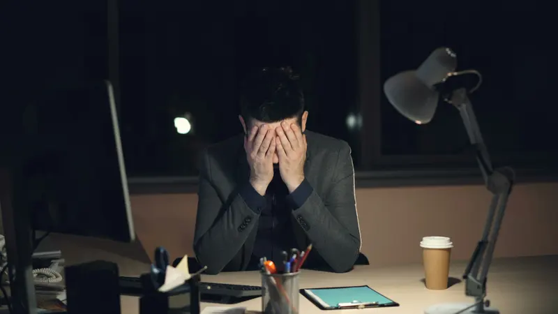 Empresário com as mãos no rosto, sentado à mesa do escritório à noite